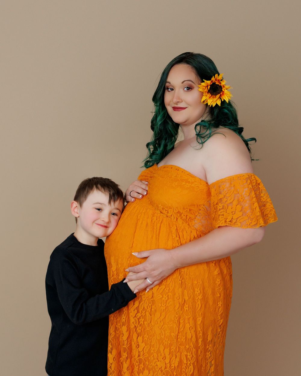 A maternity photo featuring a bright orange lace dress, sunflower accent, and a young child embracing the pregnancy bump.