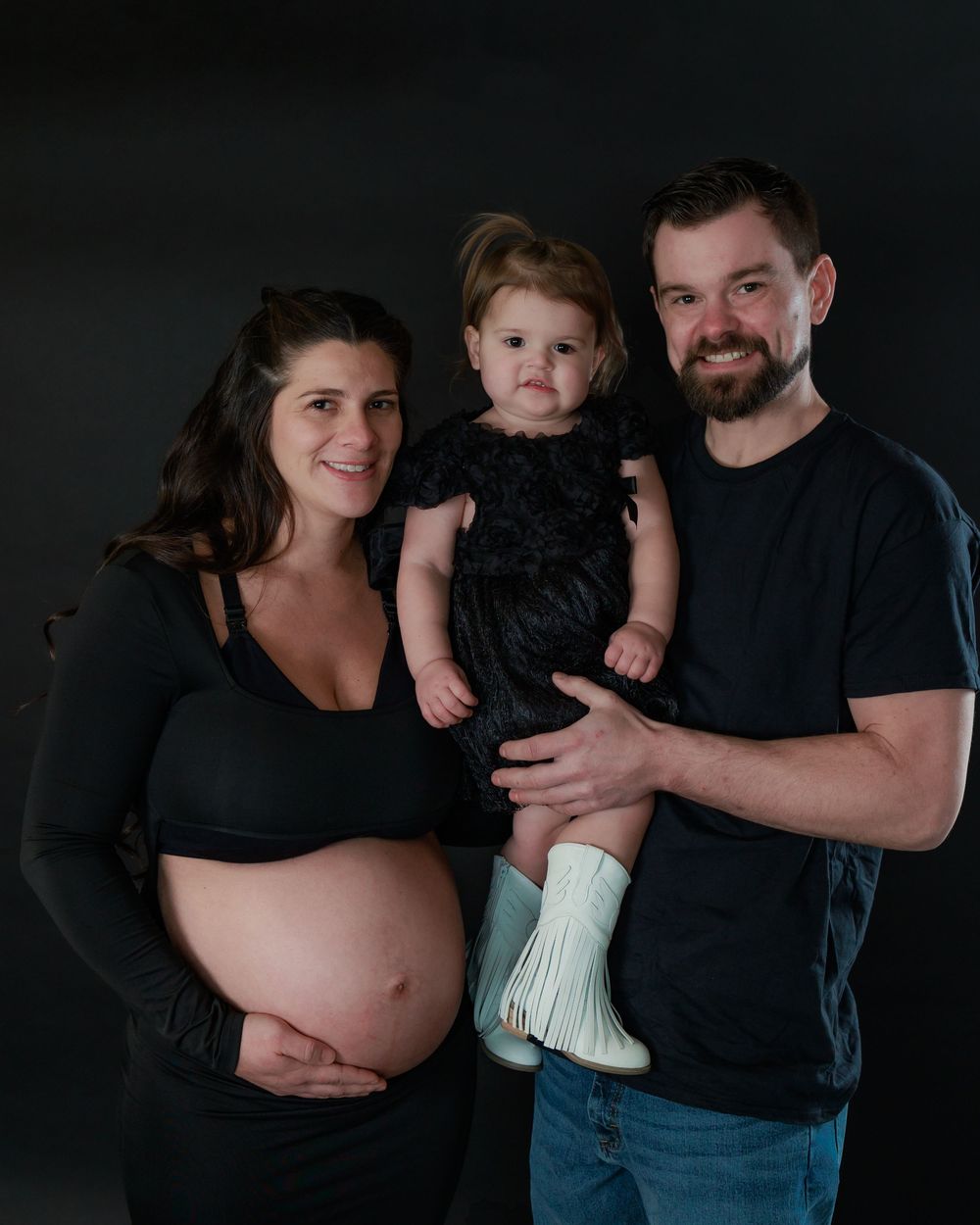 Studio portrait with black background shows an expectant couple in dark clothes with their toddler wearing a dark dress.