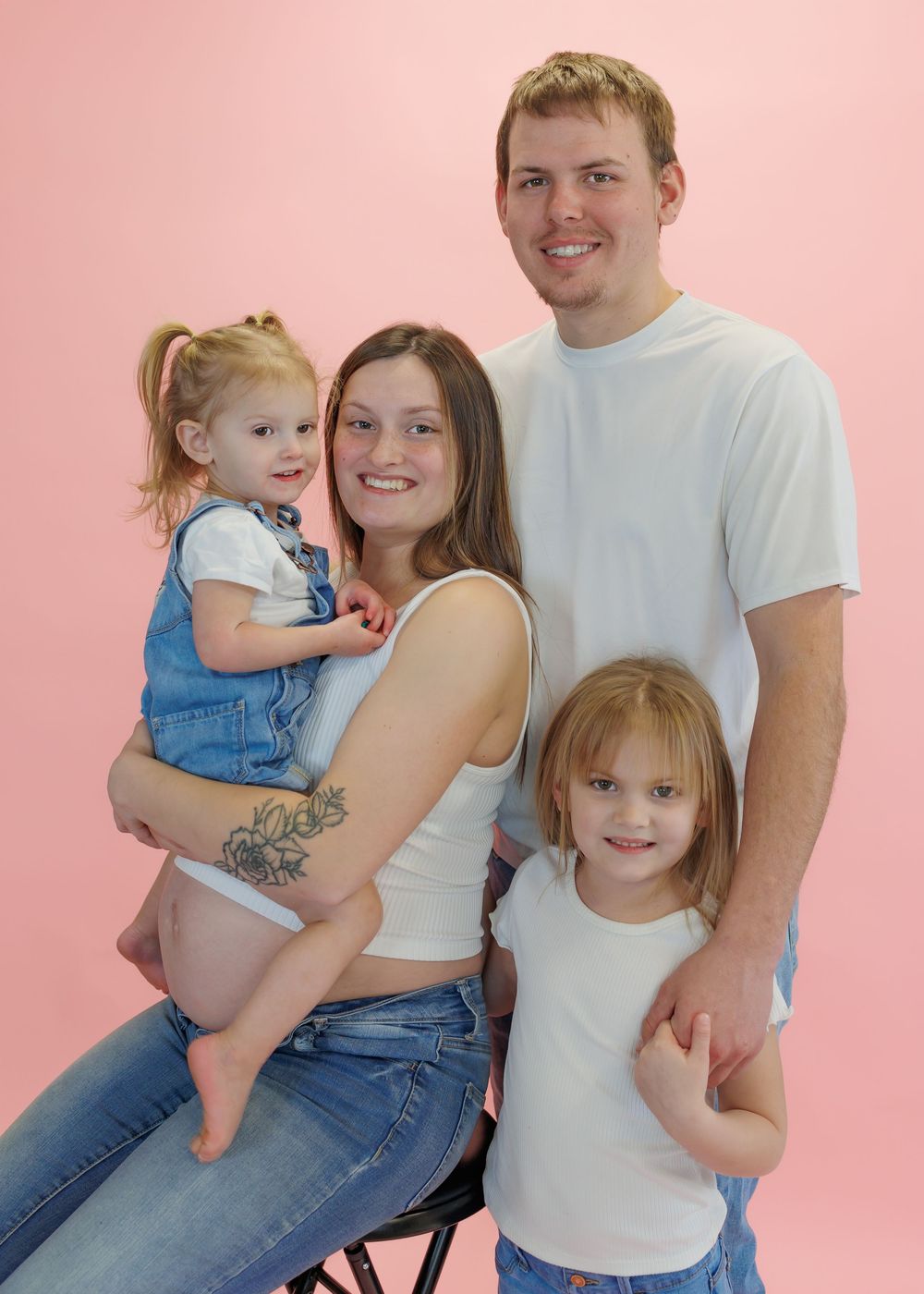 A young family of four poses together against a pink backdrop for a studio portrait.