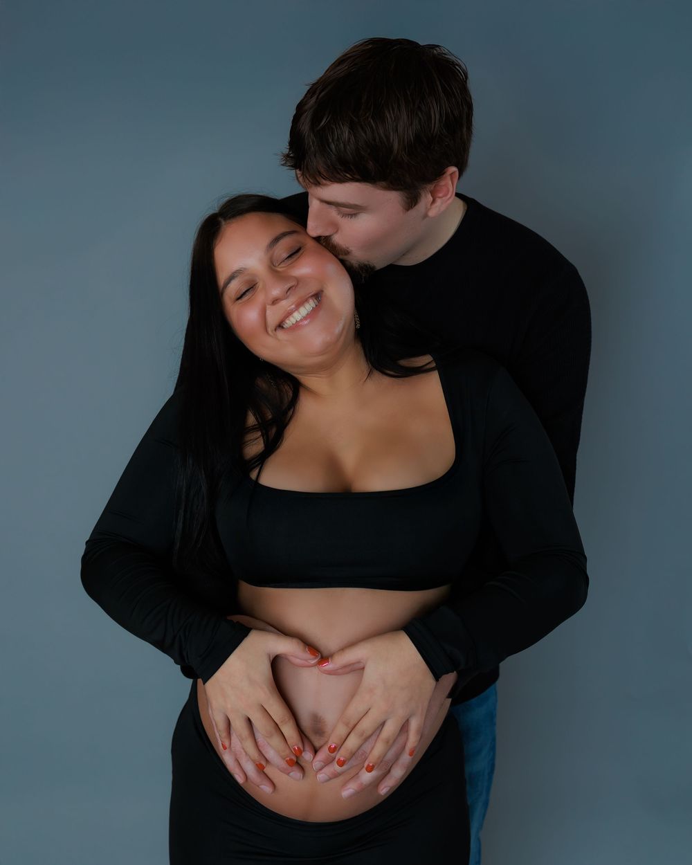 Couple in black clothing share an intimate moment while making a heart shape with their hands on a baby bump.
