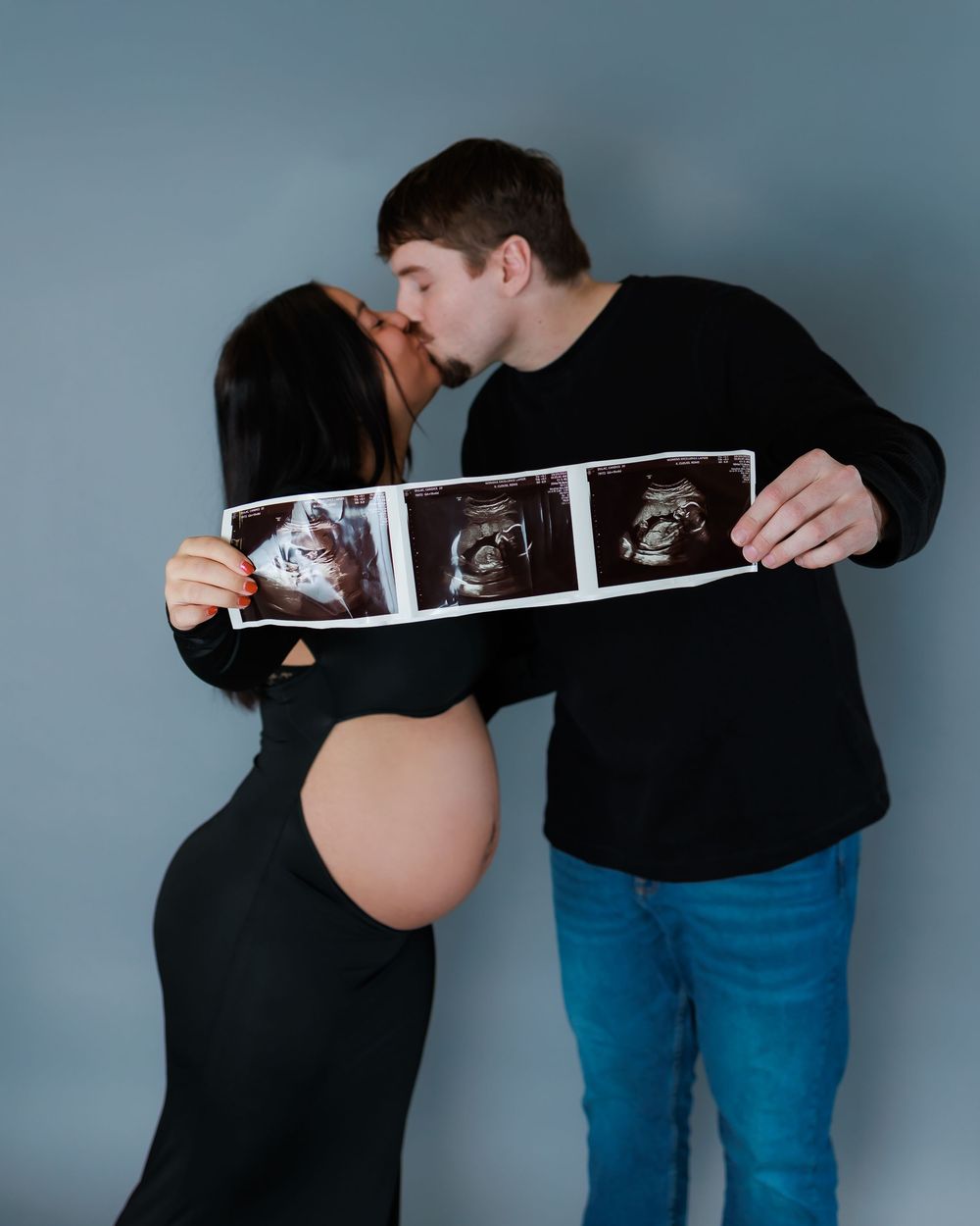 Two people in black and blue share an intimate moment while holding ultrasound photos against a blue wall.