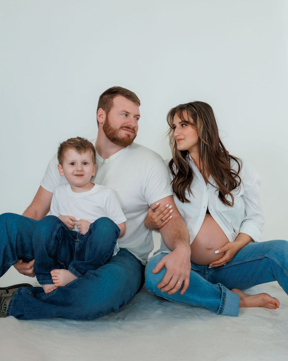 Family sitting casually together on white floor wearing jeans and light colored shirts, with woman showing bare pregnant belly.