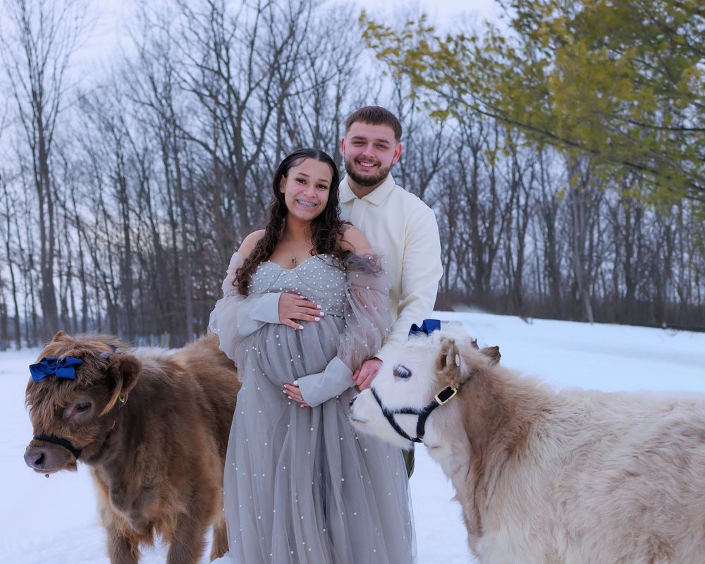 A couple poses in snowy winter scene with two dogs while wearing elegant white and gray attire for a maternity photo.