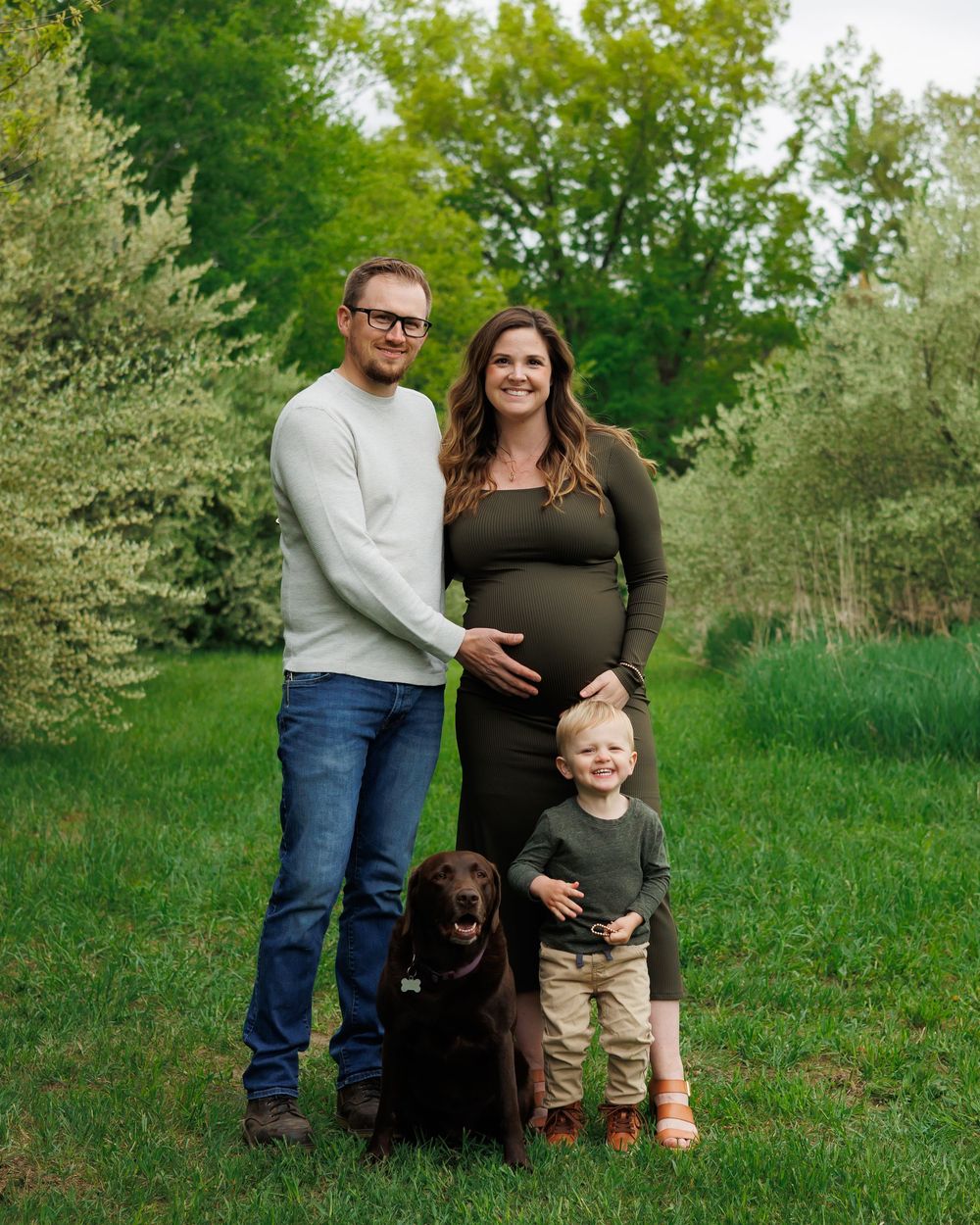 A couple and young child pose together in a grassy outdoor setting with their black Labrador retriever on a spring day.