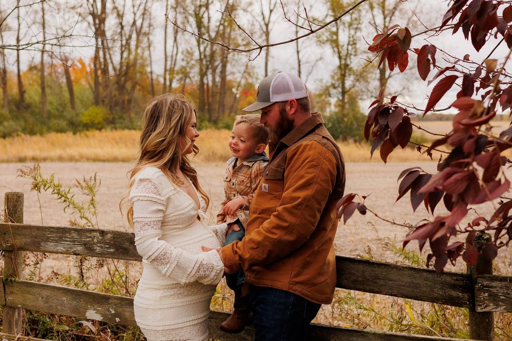 A couple in fall attire embraces by a rustic wooden fence surrounded by autumn foliage at sunset.