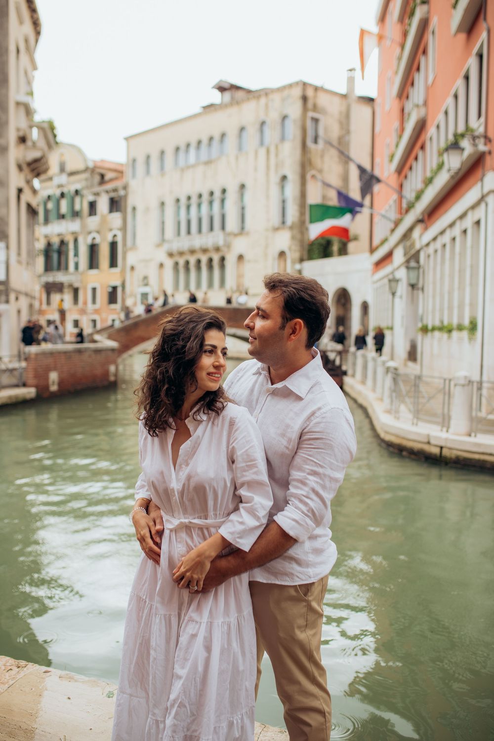 A couple shares an intimate moment along a scenic Venetian canal with colorful buildings and bridges in the background.