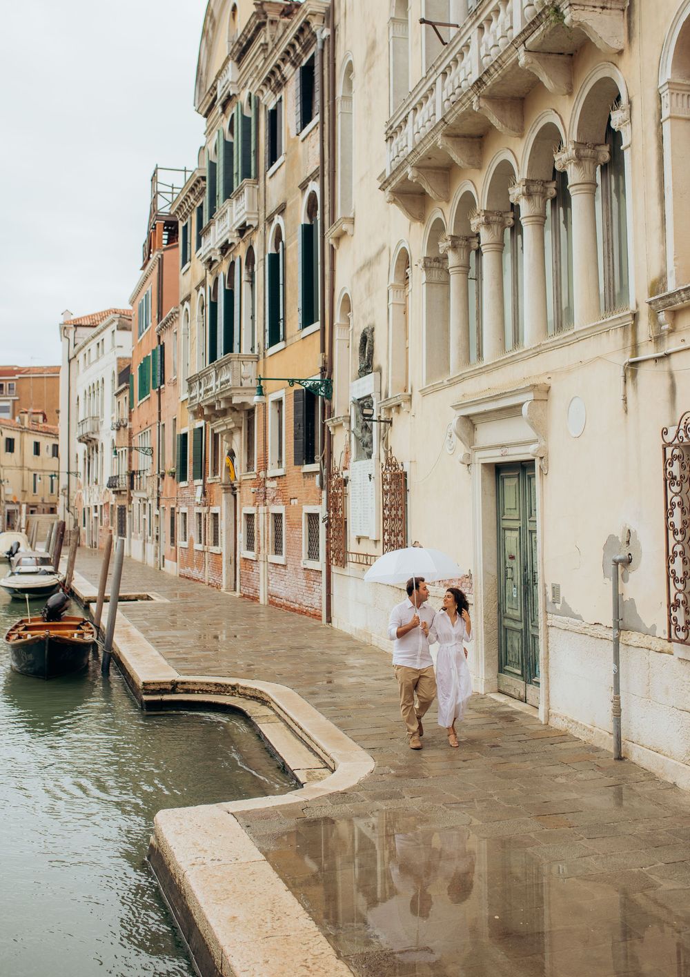 A romantic couple walking along a picturesque Venetian canal with historic architecture and arched columns.
