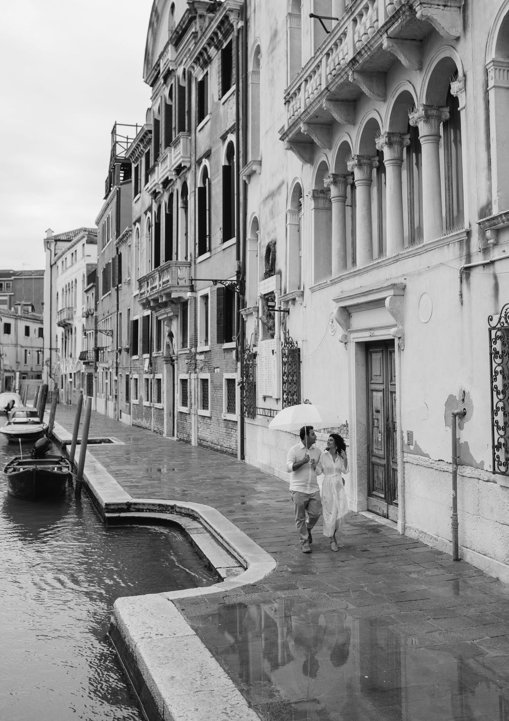 Two people walking along a scenic Venetian canal beneath white stone arches and historic buildings.