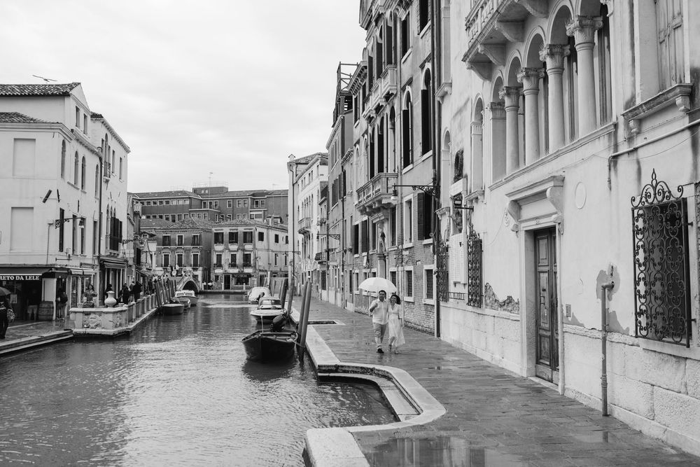 A black and white photograph of a quiet Venetian canal lined with historic buildings and traditional architecture.