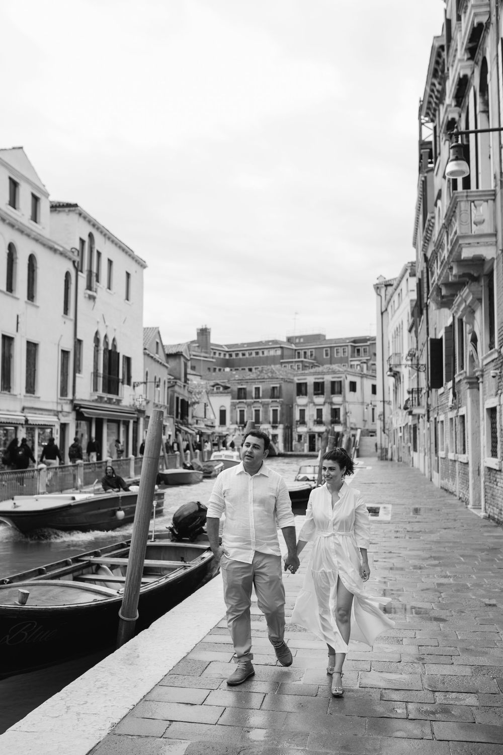 A couple walks along a Venice canal waterfront with classic Italian architecture in the background.