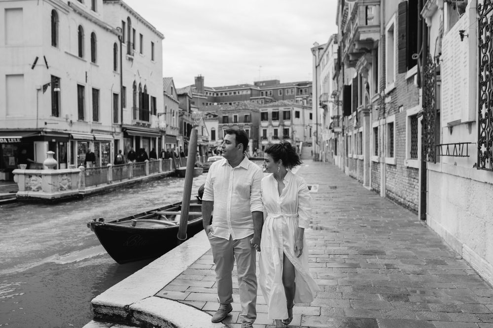 A black and white photo captures a couple walking along a quiet Venice canal street.