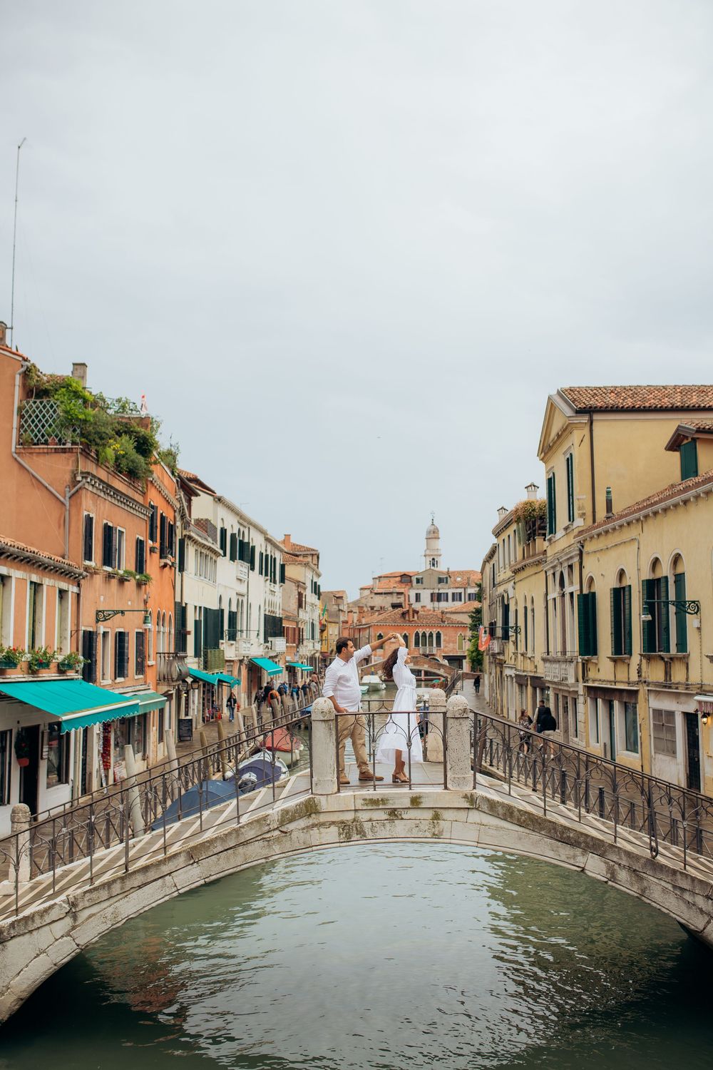 A couple stands on an arched bridge over a Venice canal with colorful buildings and architecture.