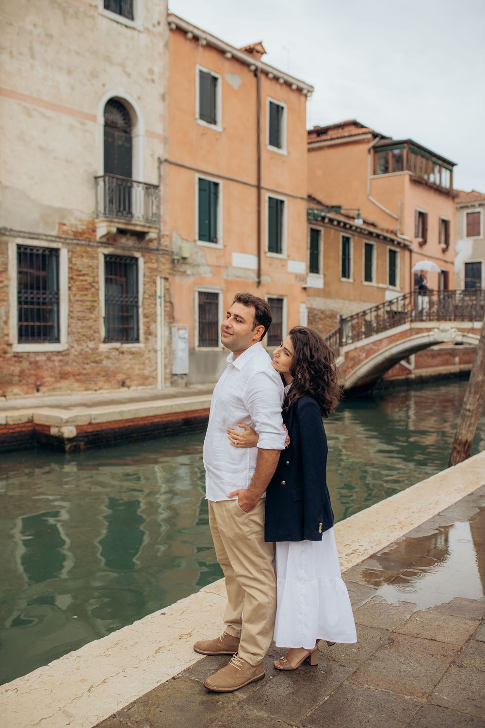 Romantic couple by a Venetian canal with colorful historic buildings and small bridge in background.