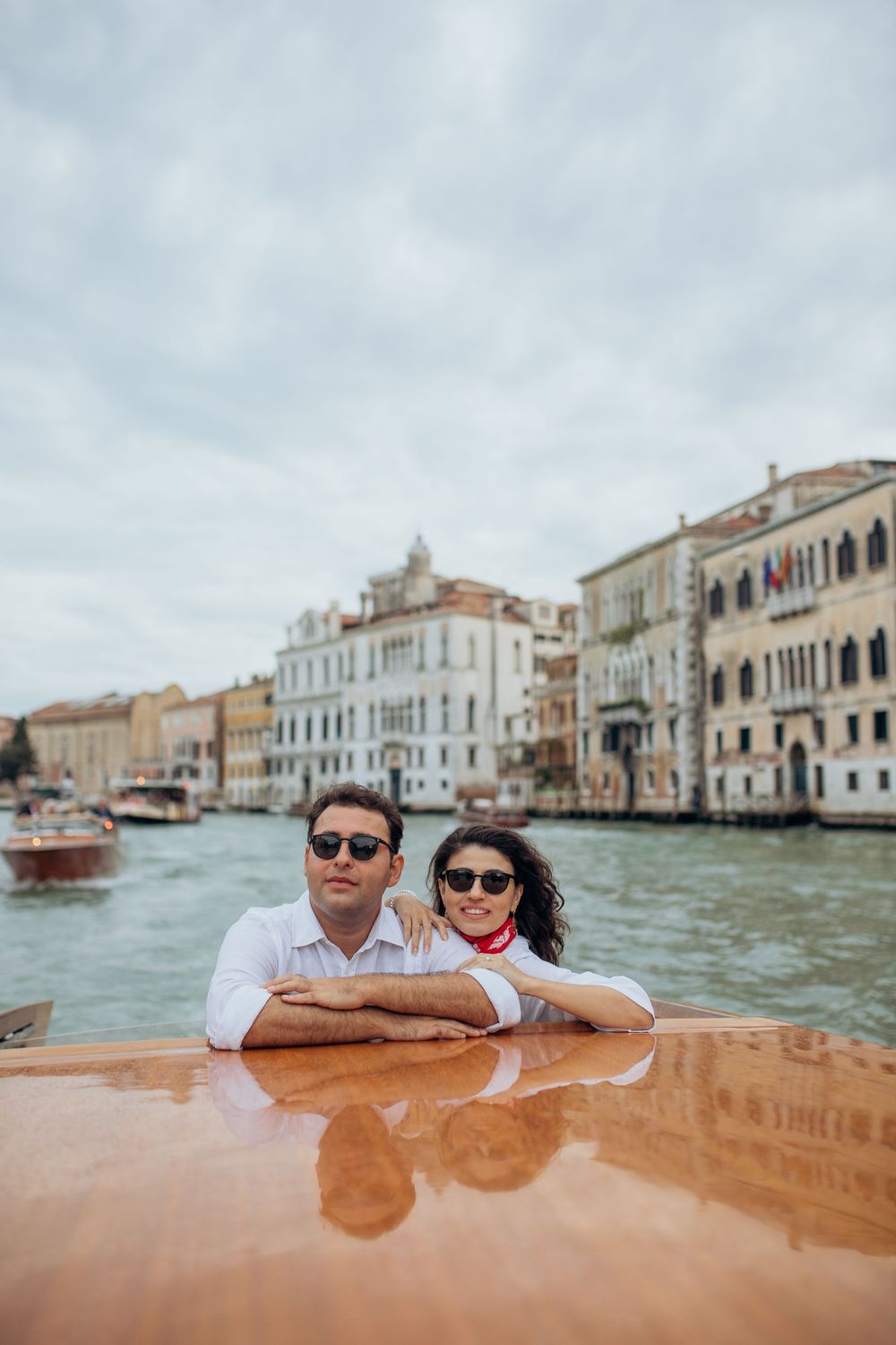 A couple enjoys a boat ride on Venice's Grand Canal, leaning against wooden railings with historic buildings behind them.