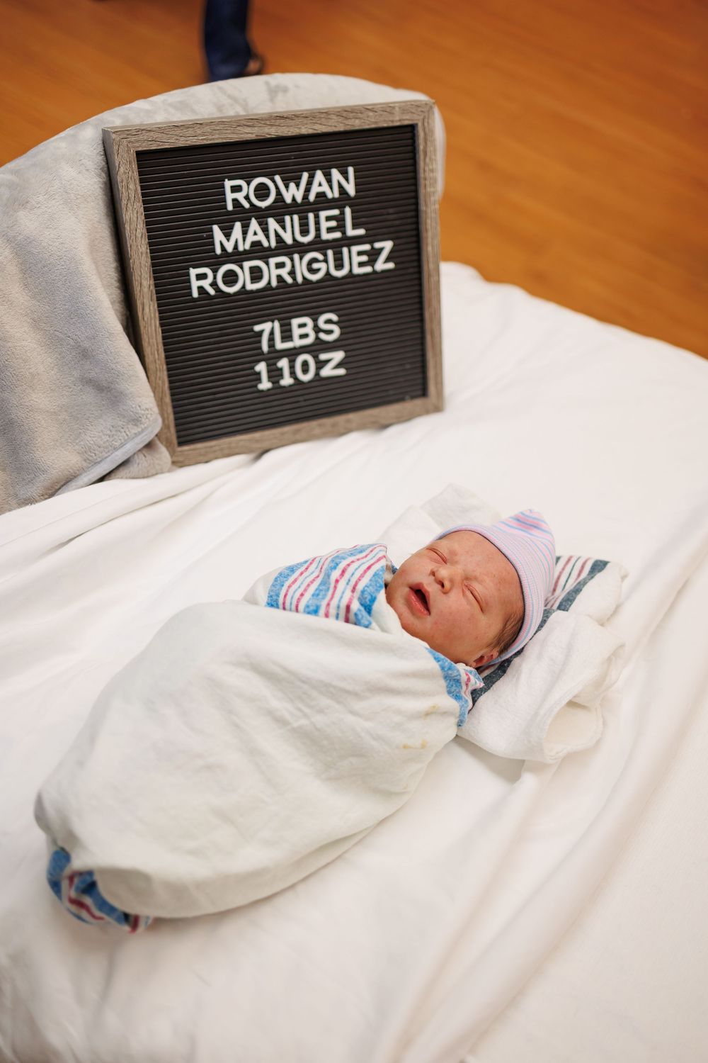 Newborn baby swaddled in white blanket next to letterboard displaying name Rowan Manuel Rodriguez and weight 7lbs 11oz.