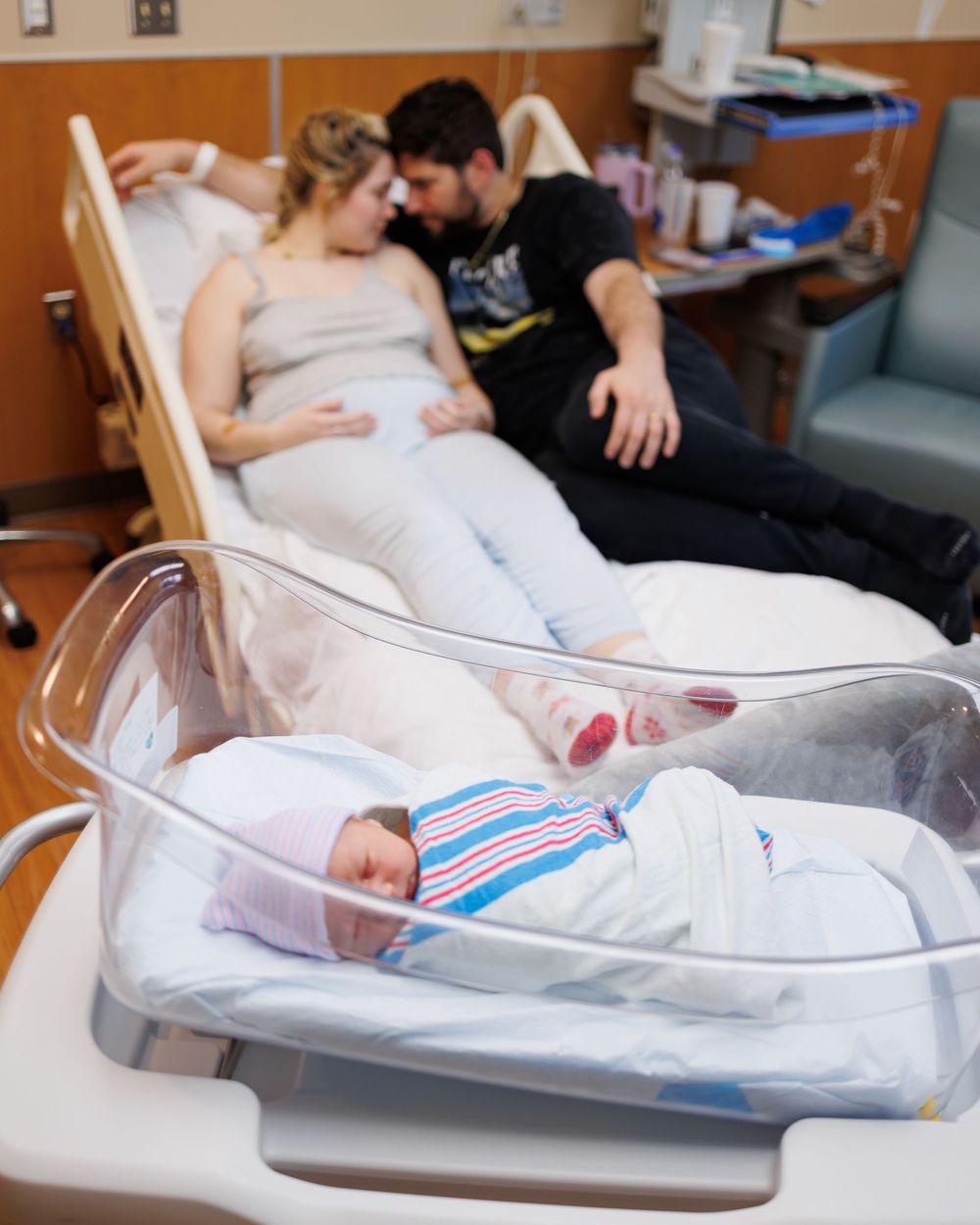A newborn baby rests in a hospital bassinet while parents share a tender moment on the bed nearby.