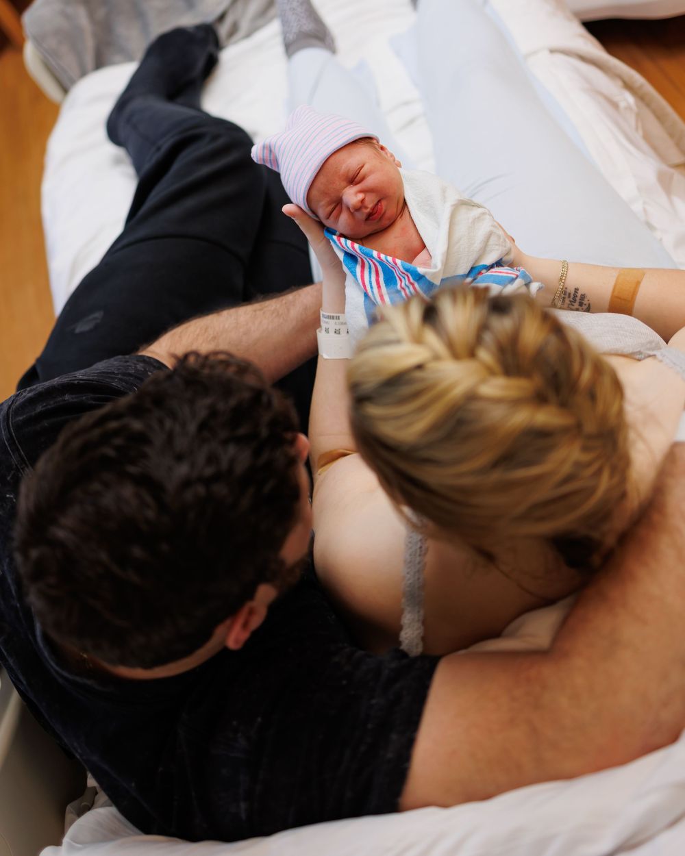 New parents lovingly gaze at their newborn baby swaddled in white while resting together on hospital bed.