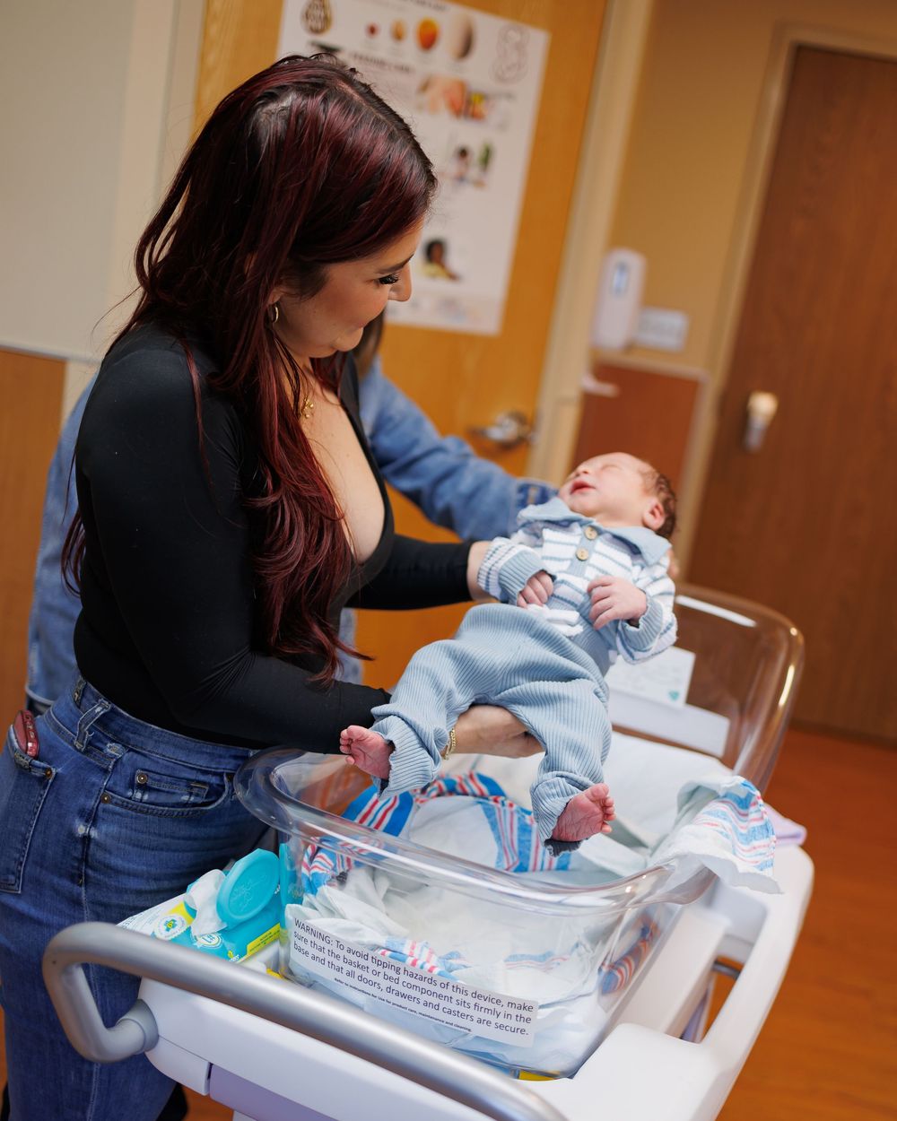 Person in black top and jeans tending to a newborn baby lying in a hospital bassinet during medical care.