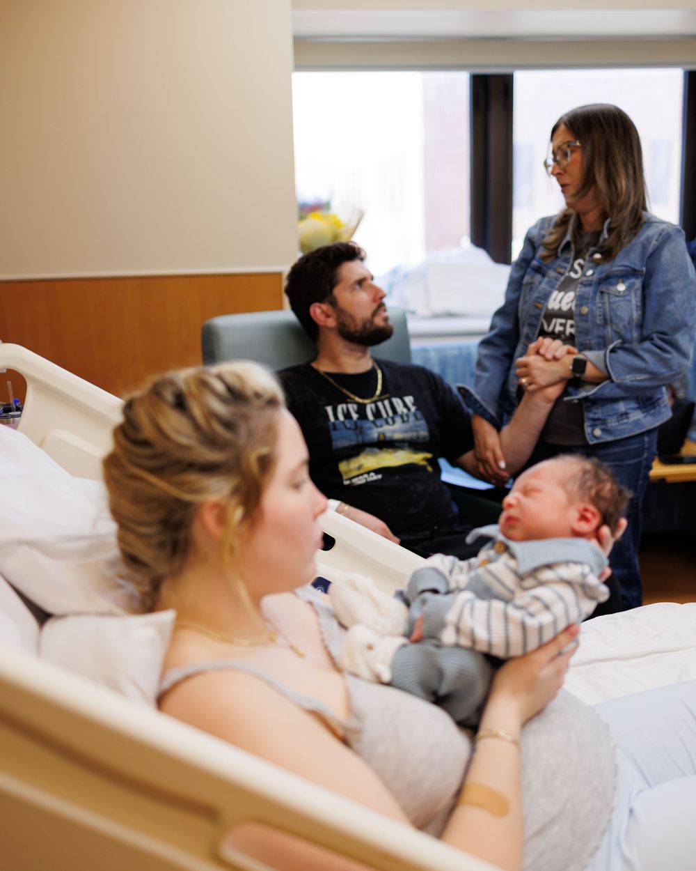 Family together in hospital room as woman rests in bed holding newborn baby while others visit and support.