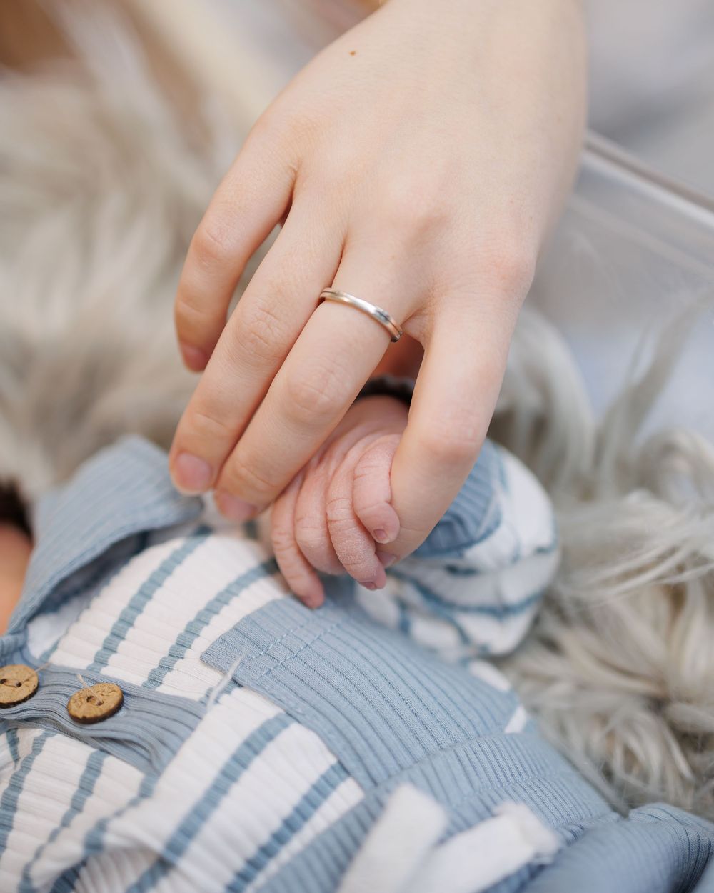 A delicate hand wearing an elegant ring gently holds a tiny infant's hand against a soft blue and white striped fabric.