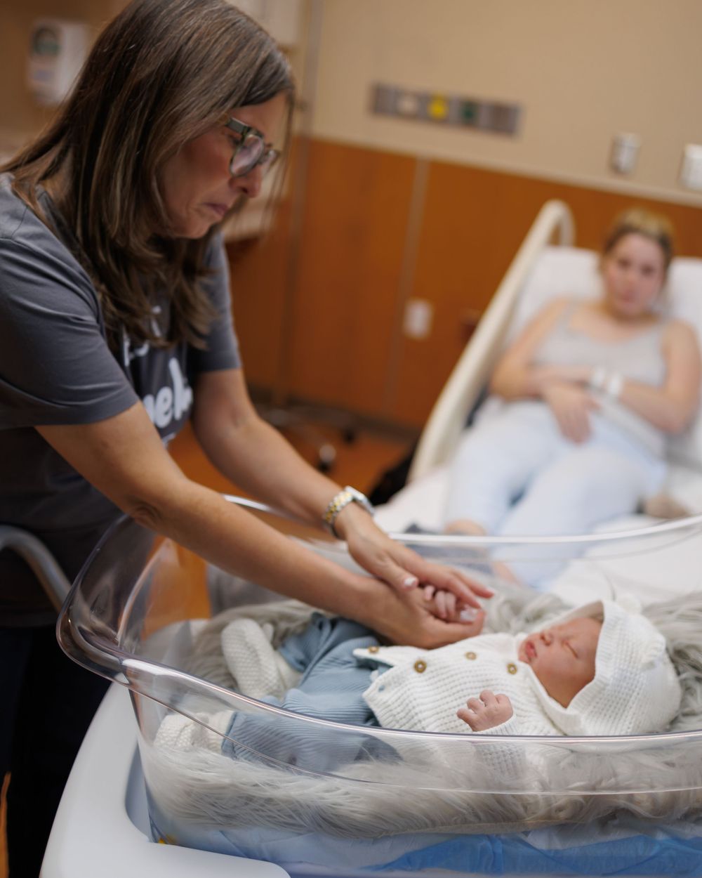 Medical professional checking on newborn baby in bassinet while parent rests in hospital bed.