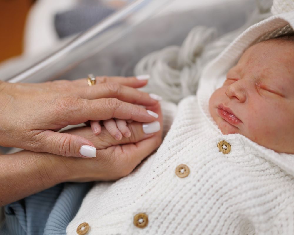 A sleeping baby in a white knit sweater with brown buttons grasps a caring hand.
