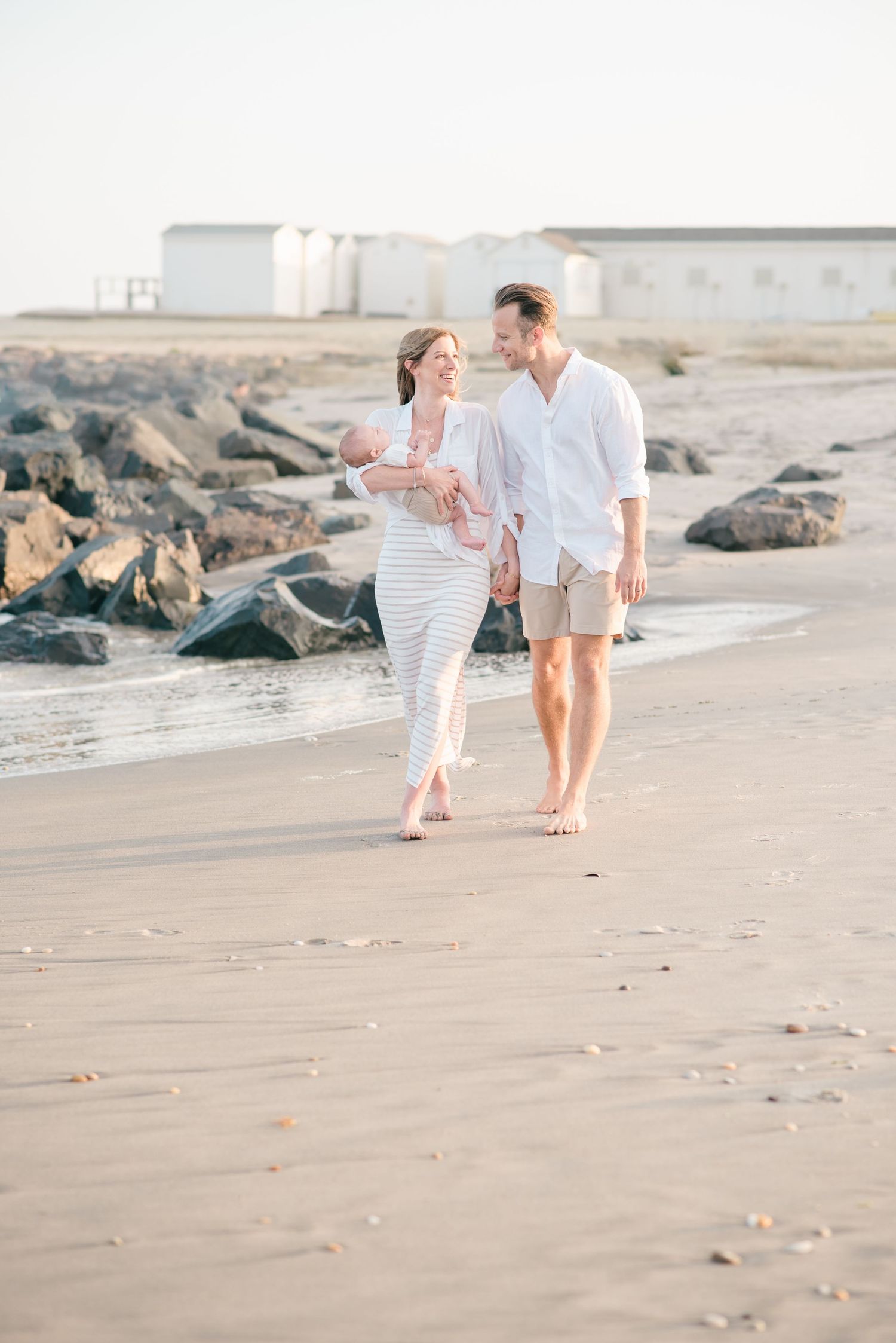 A man and woman walk down the beach holding hands smiling at each other while the woman holds her son in her right arm.