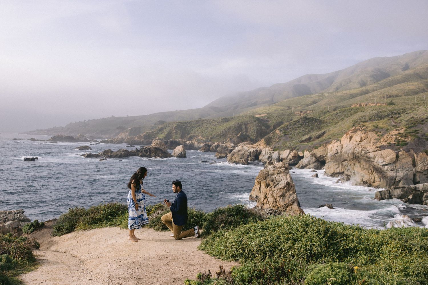 Two people stand on a cliff overlooking the rugged Big Sur coastline with waves crashing against rocky shores below.