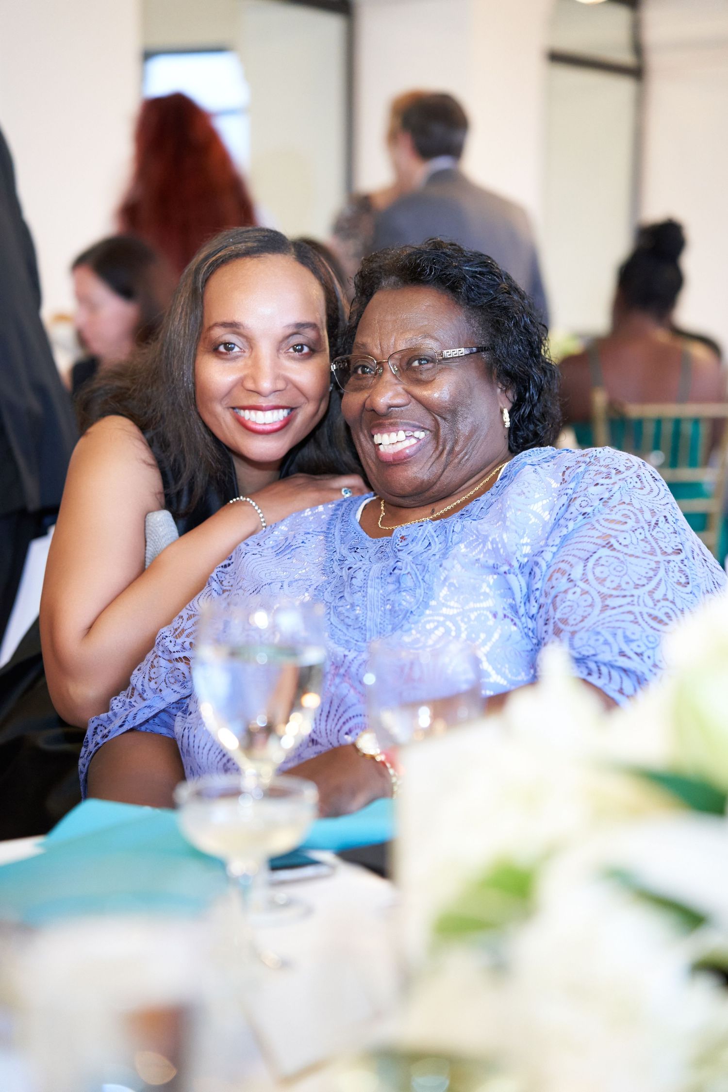 Two women smile warmly together at an elegant event, wearing coordinating blue lace outfits.
