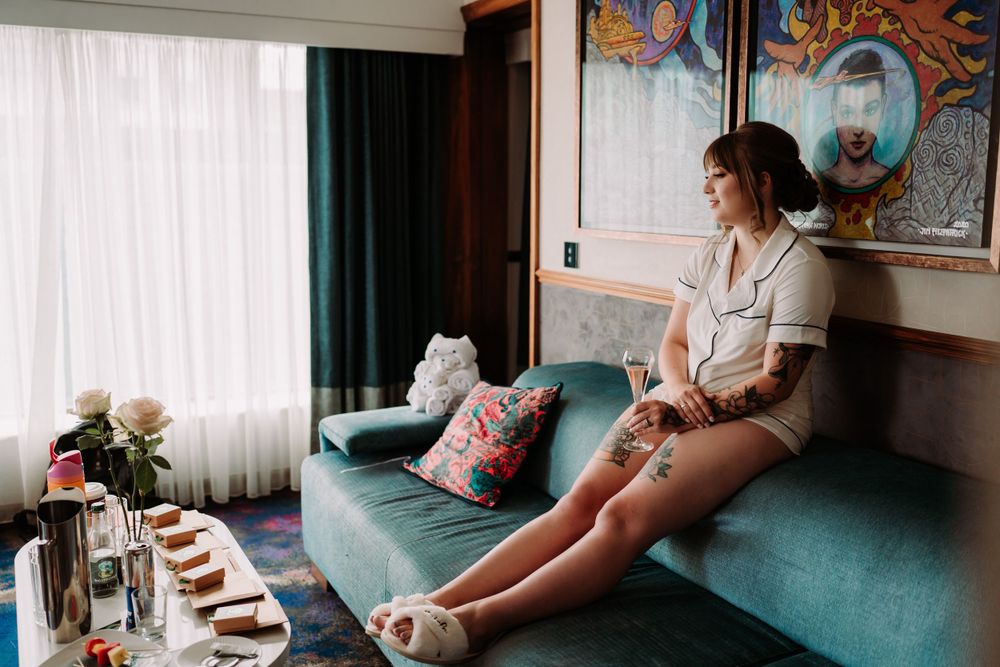 A person in white sits on a teal couch near floral arrangements and throw pillows in a light-filled room.