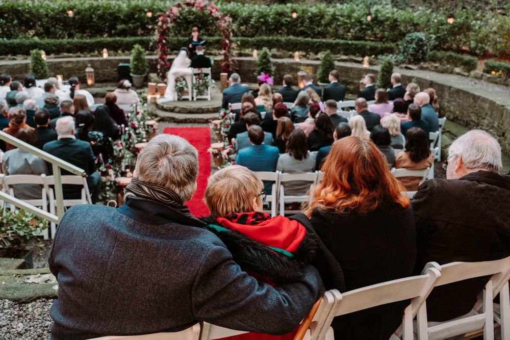 Guests seated on white chairs attend an outdoor garden wedding ceremony with a red carpet aisle and flowering archway.