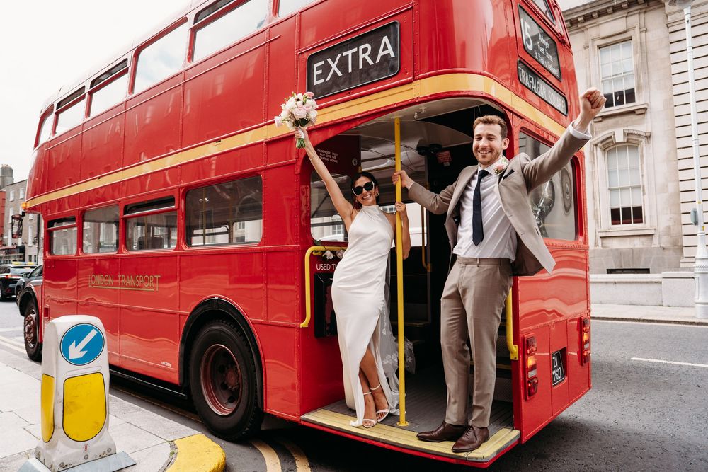 A celebratory couple poses on a classic red London double-decker bus on their wedding day.