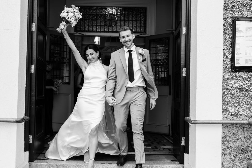 A joyful couple exits a building after their wedding ceremony with the bride holding up her bouquet.