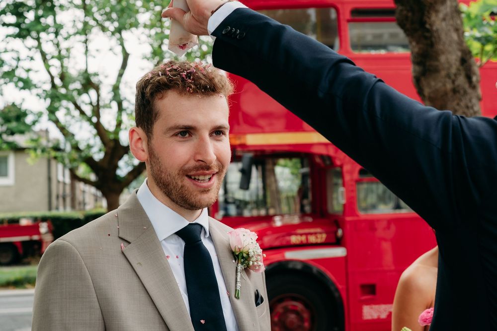 A joyful wedding moment captured in front of a vintage red double decker bus with trees in the background.