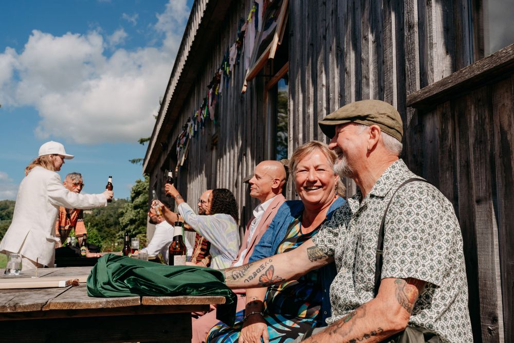 People enjoying a sunny afternoon socializing on wooden benches outside a rustic barn building.
