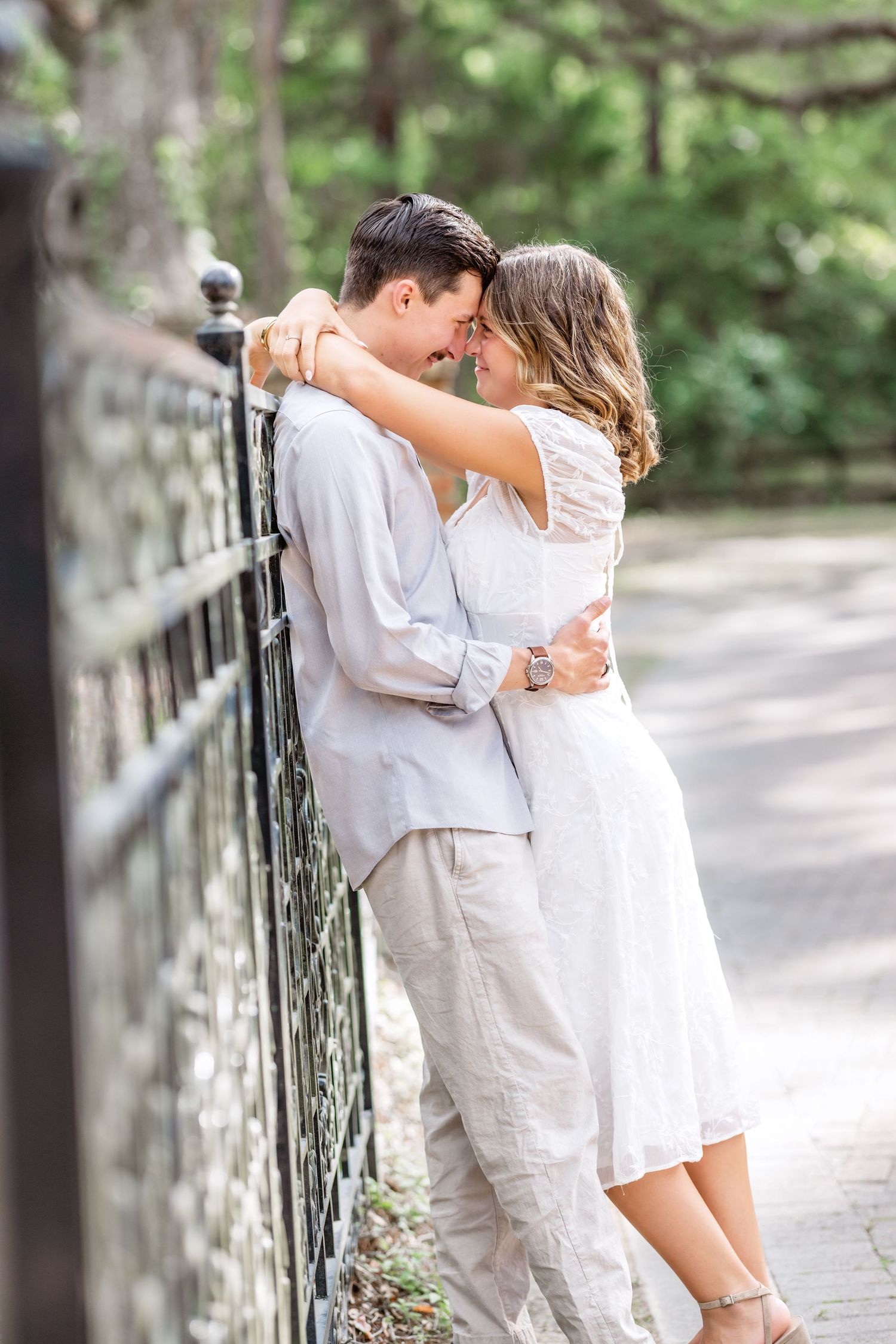 A romantic couple shares a tender embrace against a wrought iron railing in a park setting wearing white summer outfits.