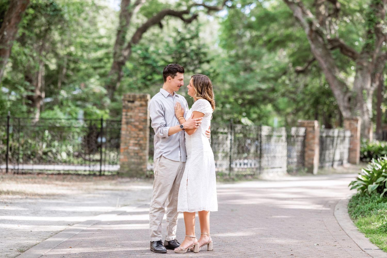 Two people share an intimate moment together on a sunlit path in a lush park setting surrounded by trees and greenery.