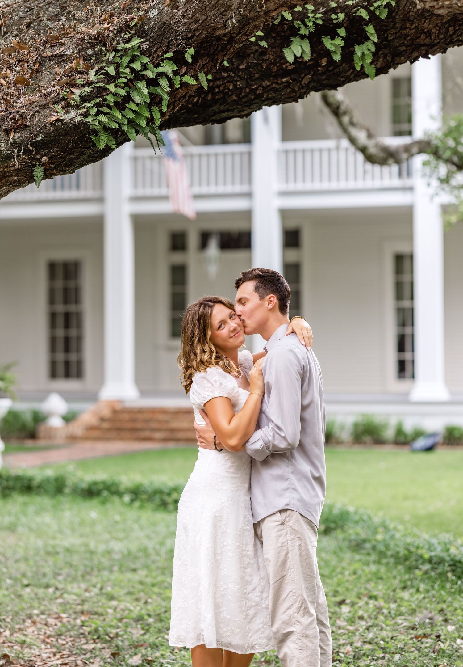 A couple shares a romantic moment in front of a white antebellum style mansion with large columns and a wraparound porch.