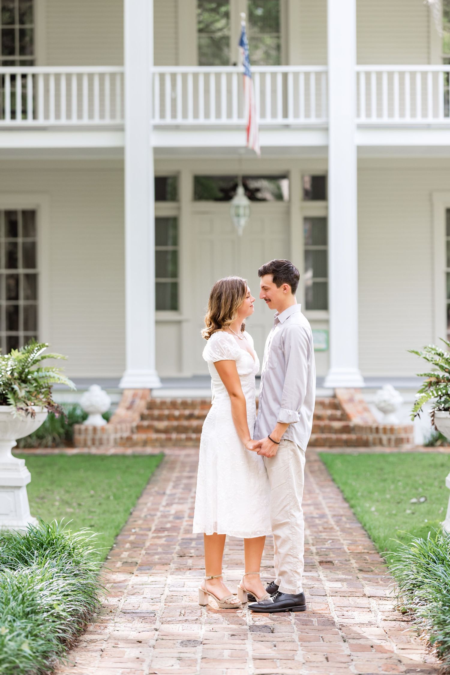 A couple in white and light colored clothing stand together on a brick pathway in front of a colonial-style porch.