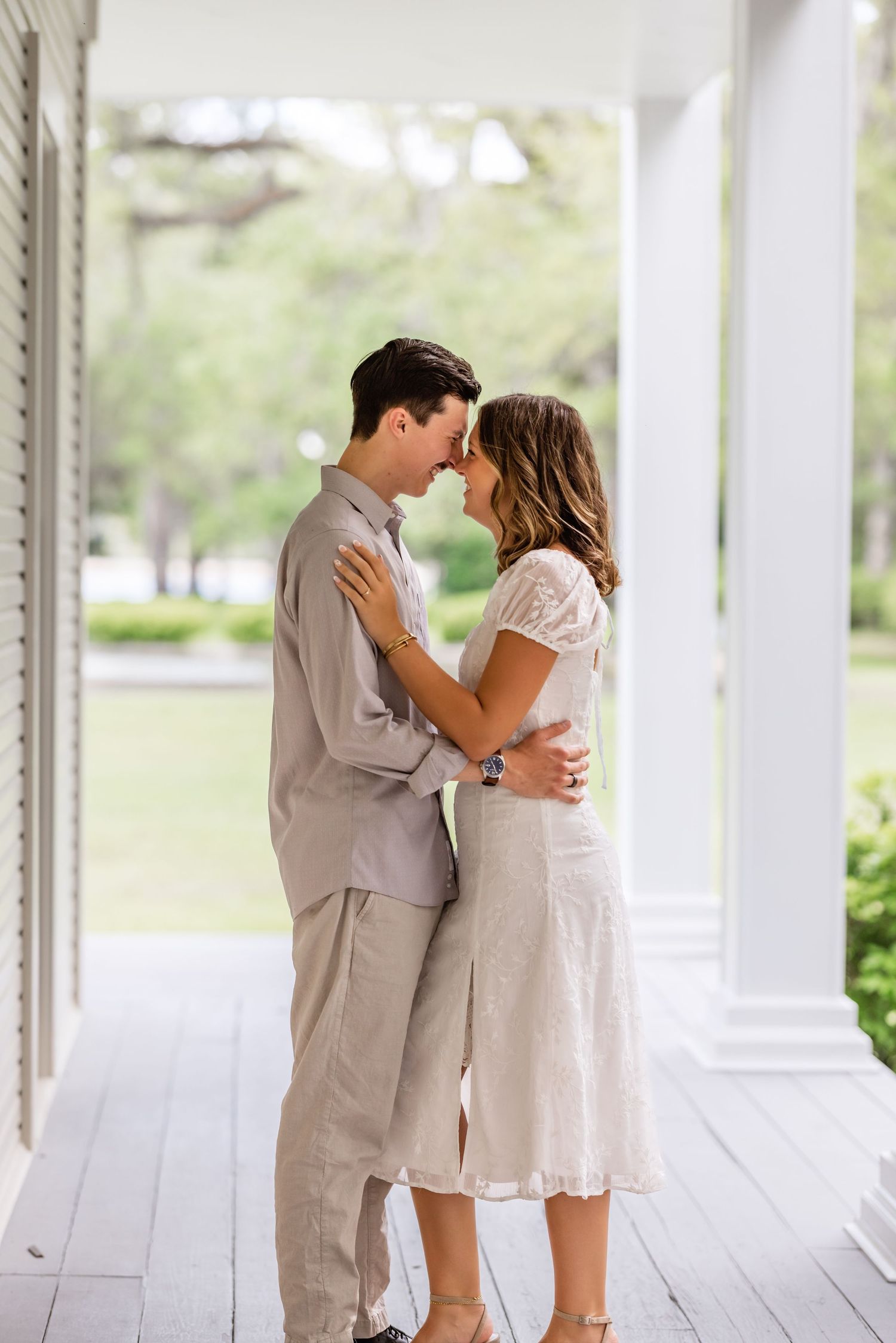 A romantic couple shares an intimate moment on a bright white porch in light colored outfits.