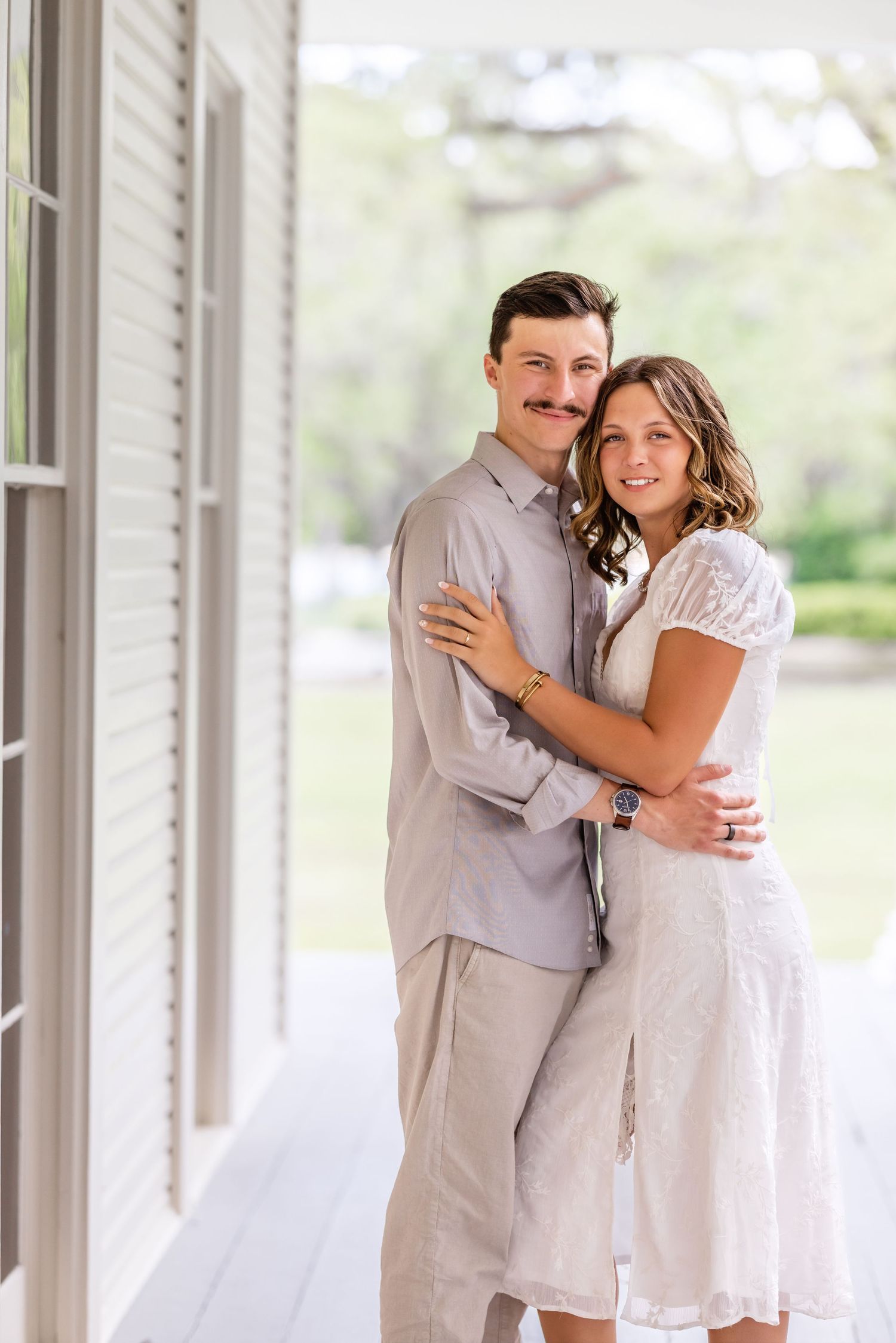 A romantic couple in coordinated light-colored outfits embrace on a white porch on a bright sunny day.