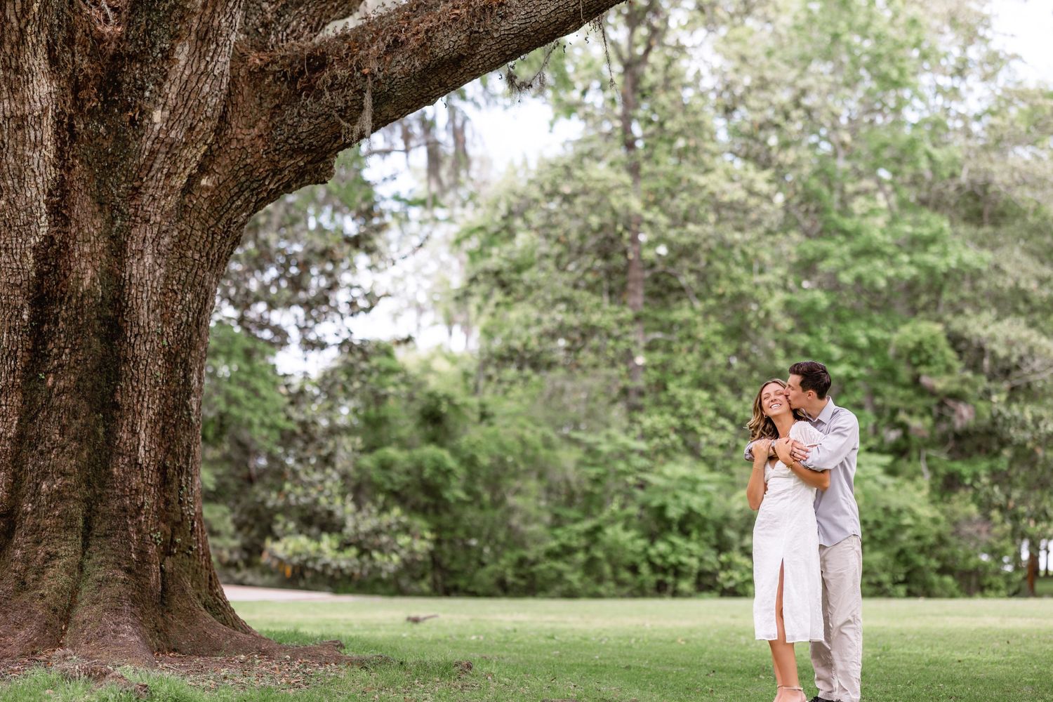 A couple embraces under a majestic oak tree in a lush green garden setting during a romantic outdoor moment.
