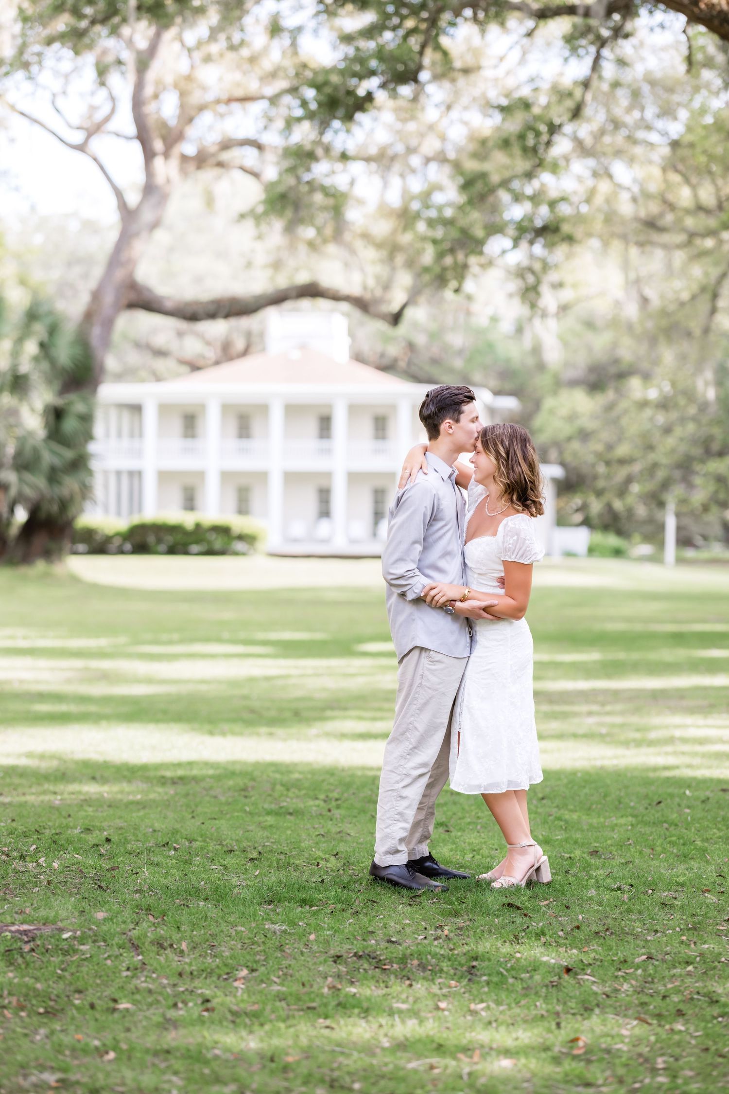 A couple shares an intimate moment on a lawn with a grand white Southern mansion visible in the background.