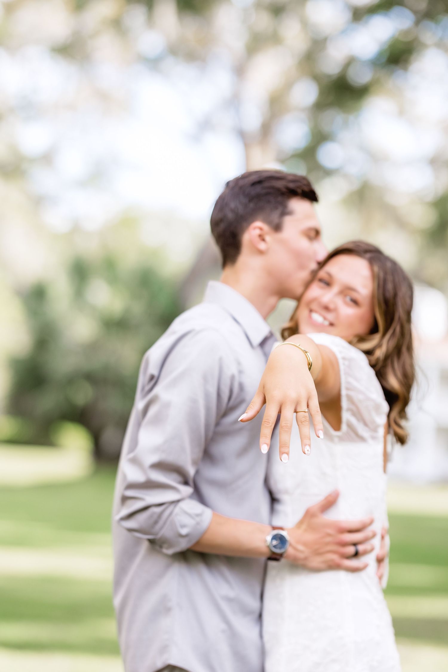 Engaged couple embraces in sunlit park as she displays engagement ring while sharing intimate moment together.