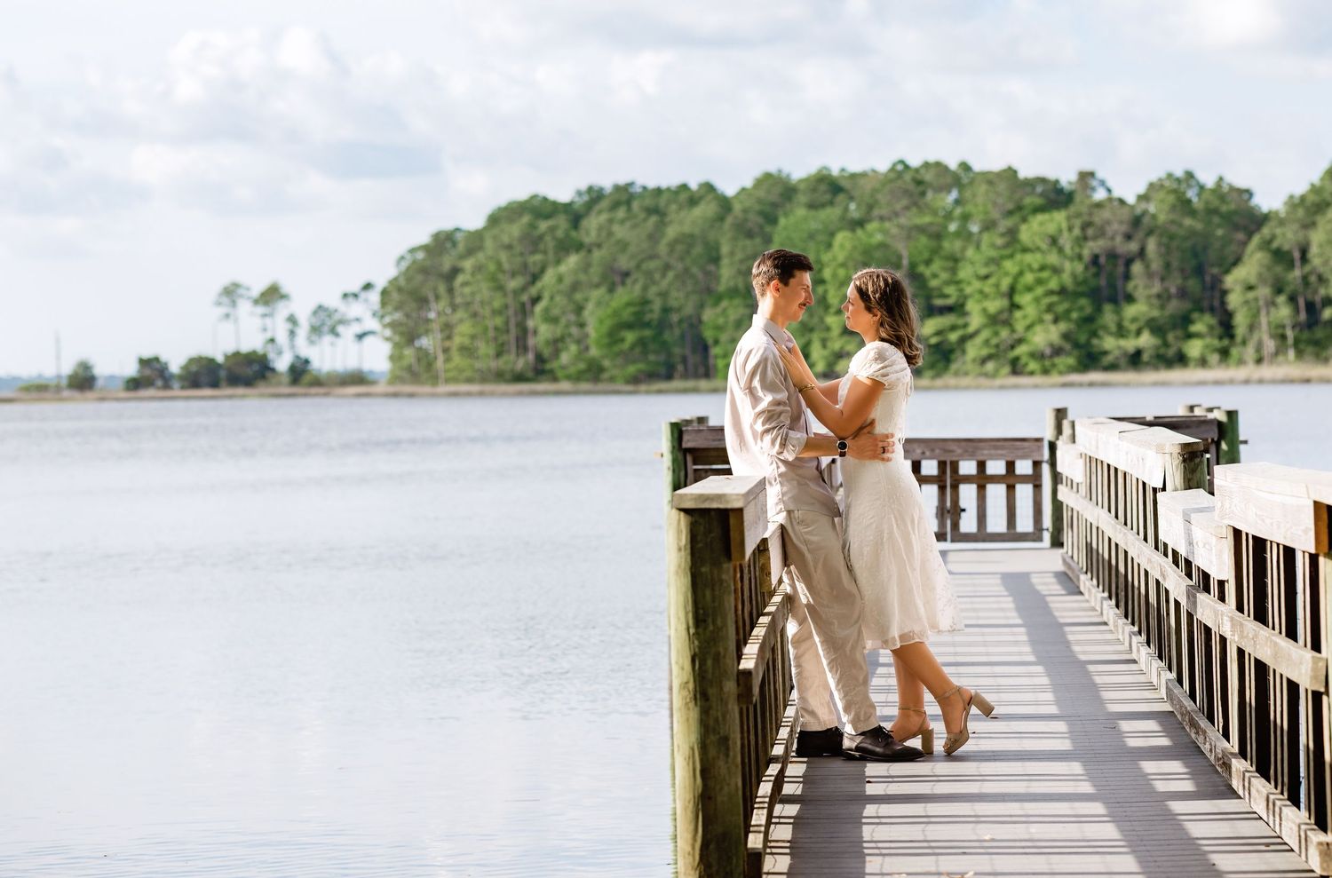 A couple stands together on a wooden pier overlooking a serene lake surrounded by trees on a bright sunny day.