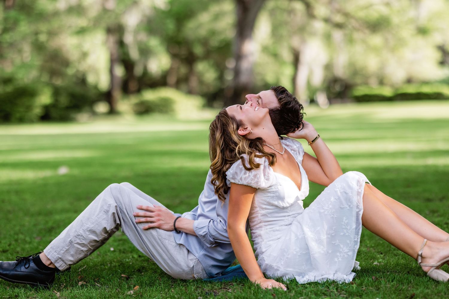 Two people in casual attire sitting and embracing on green grass in a sunny park setting.