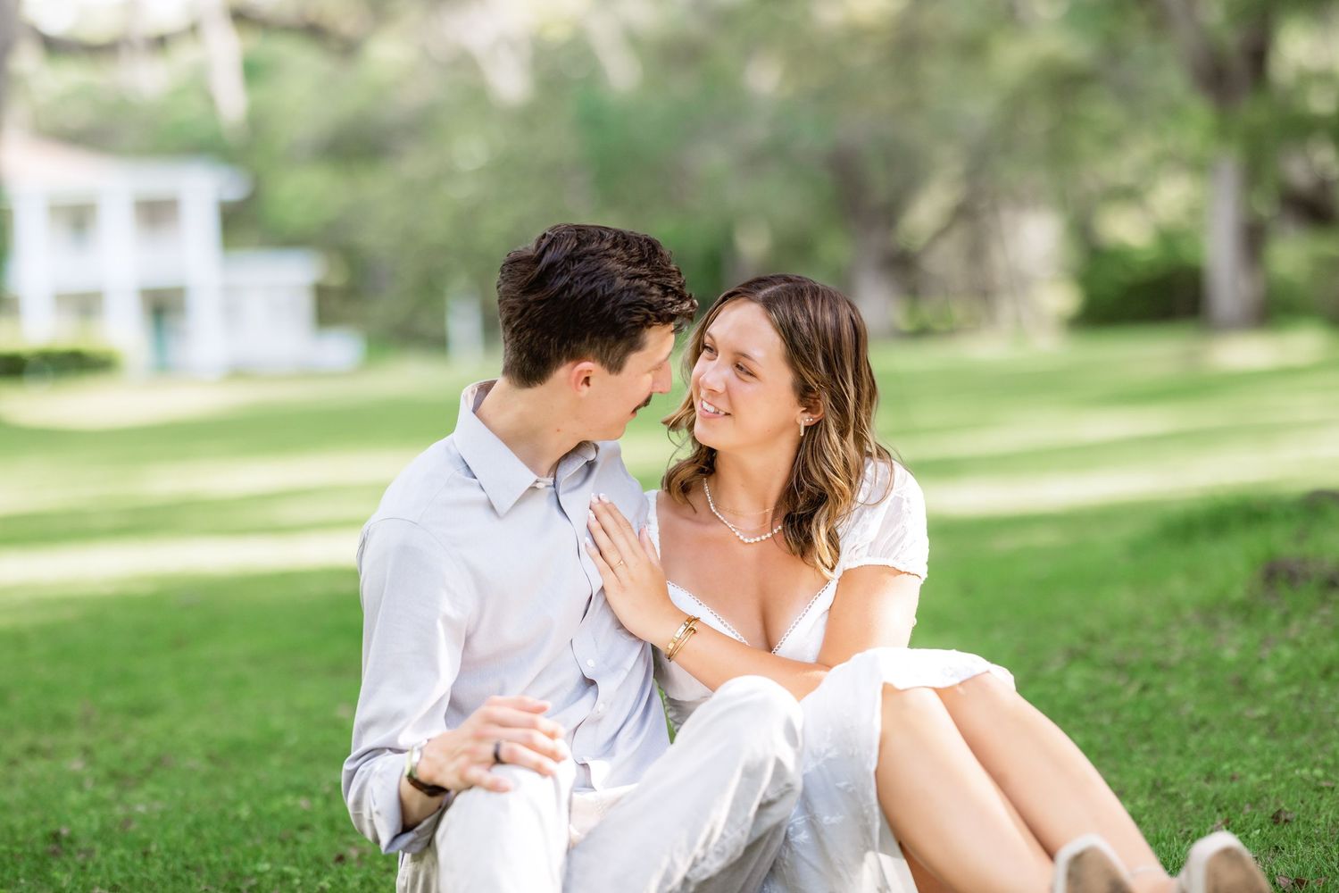 A couple in white sits together intimately on grass in a sunlit park setting.
