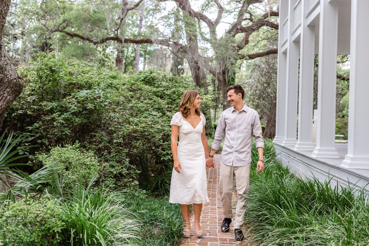 Couple walking together on garden path beside lush greenery and white columned building on sunny day.