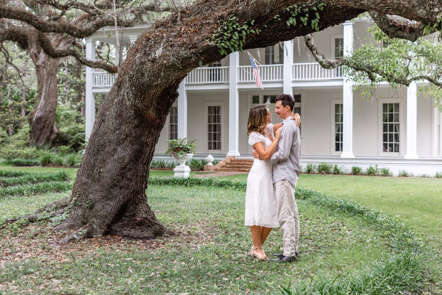 A romantic moment captured under a large oak tree in front of a white colonial style mansion house.