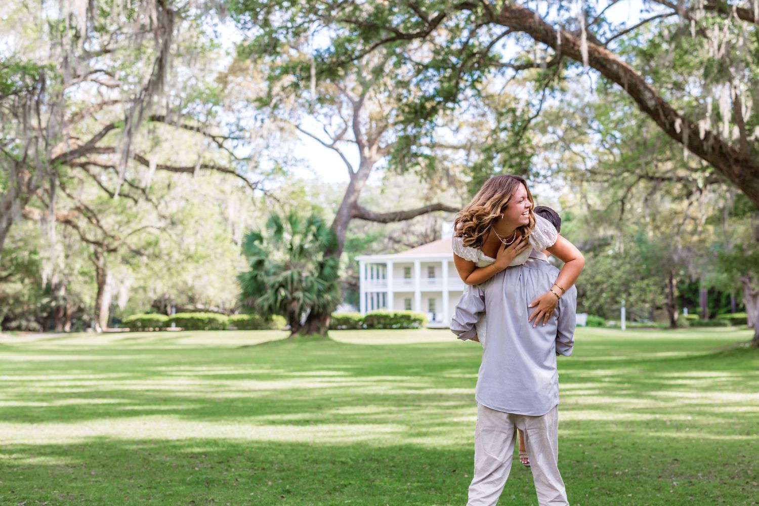 Someone in white embraces a fluffy dog while standing on a lush lawn in front of a stately Southern plantation home.